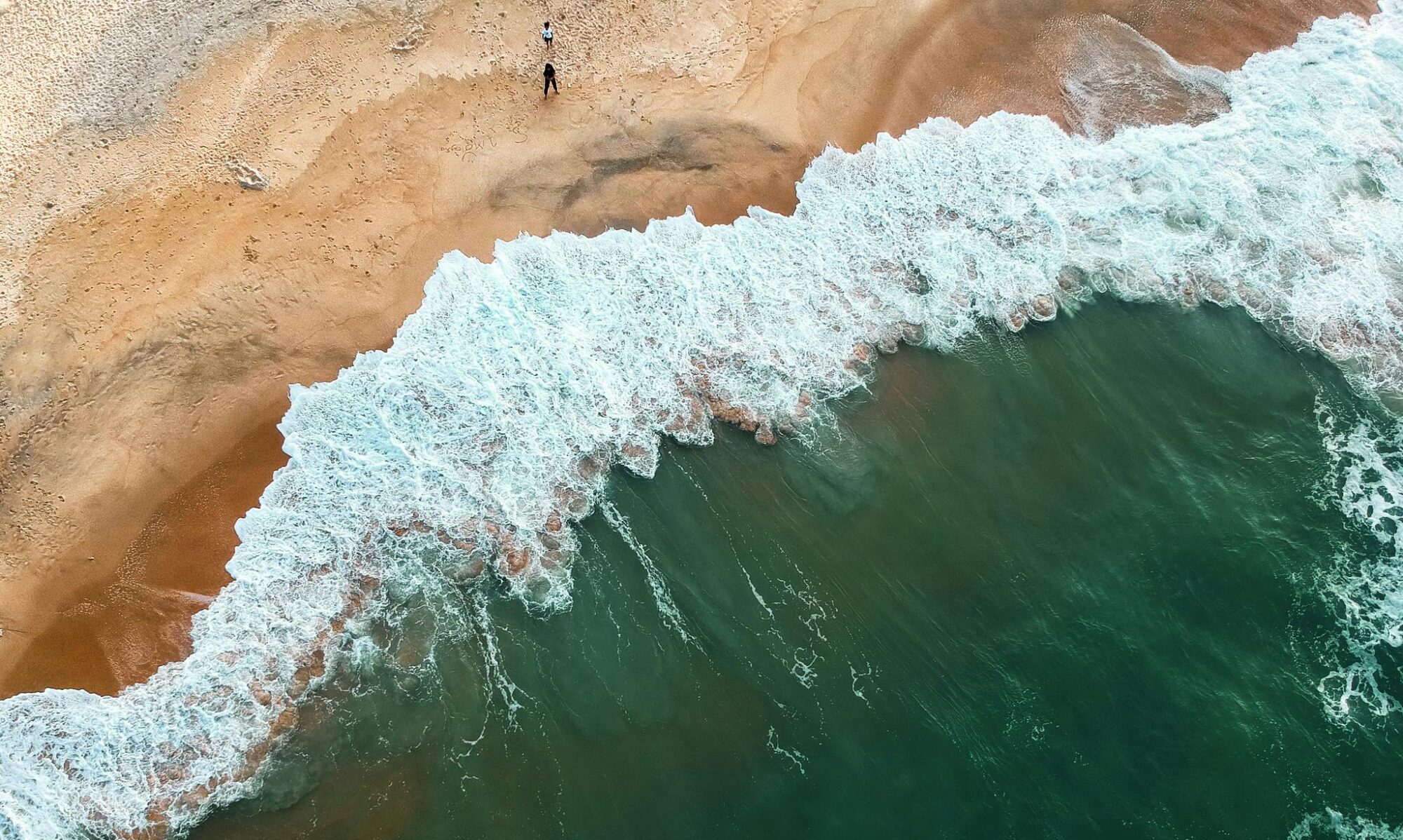 Aerial shot of ocean waves crashing onto a sandy beach with two people in the distance.